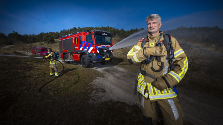 Brandweerman in beschermpak spuit water; op de achtergrond een rode brandweerwagen met “Brandweer Schoorl”