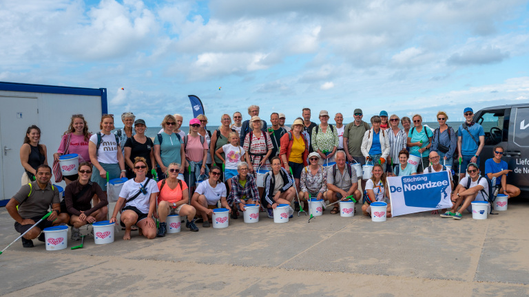 Groepsfoto van strandopruimers met emmers en grijpers; banner met tekst Stichting De Noordzee op de achtergrond