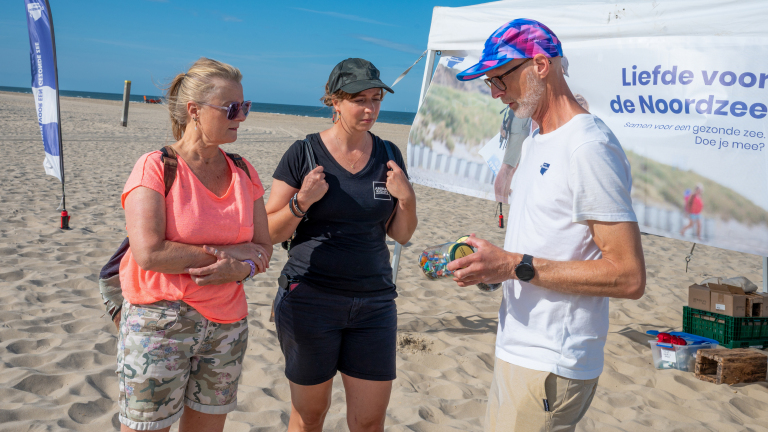 Drie mensen praten op het strand bij een stand met banner “Liefde voor de Noordzee”, terwijl een man een potje toont