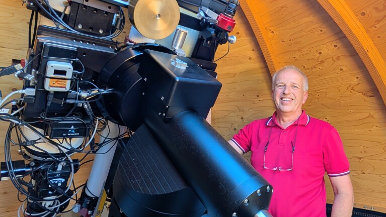 Man in rood poloshirt naast een grote telescoop met camera’s en kabels in een houten sterrenwacht