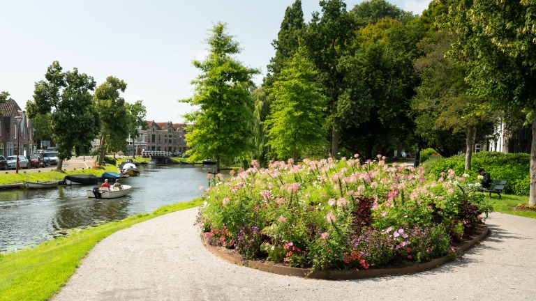 Bloemenperk langs een kanaal met wandelpad, bomen en een kleine boot; in de verte huizen en een brug