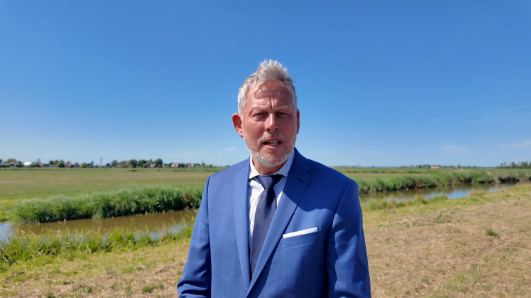 Man in blauw pak en stropdas staat buiten bij een sloot in een groen polderlandschap onder blauwe lucht