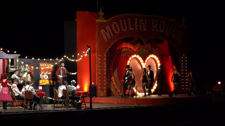 Avondvoorstelling in een rode Moulin Rouge-achtige cabaretzaal met dansers op het podium en publiek aan tafeltjes