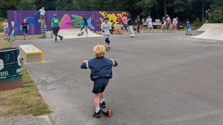 Kind op een step in een skatepark, met skaters en toeschouwers voor een muur met kleurrijke graffiti