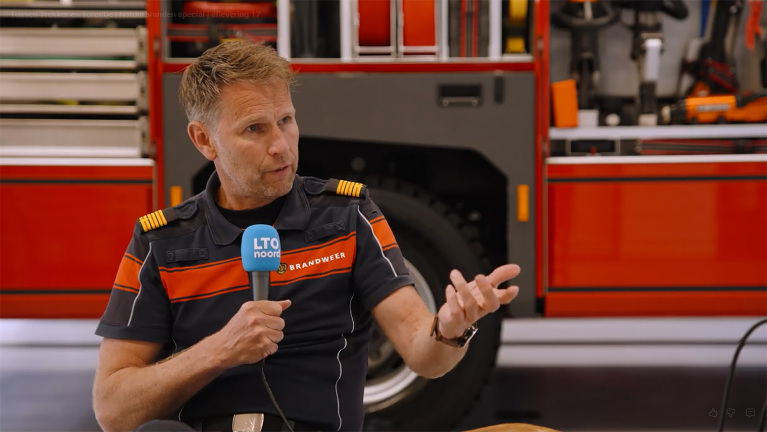 Brandweerman in uniform geeft interview met LTO Noord-microfoon, met brandweerwagen op de achtergrond