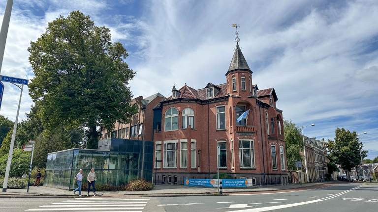 Rood bakstenen gebouw met torentje en windvaan naast glazen aanbouw, met grote boom en passerende voetgangers