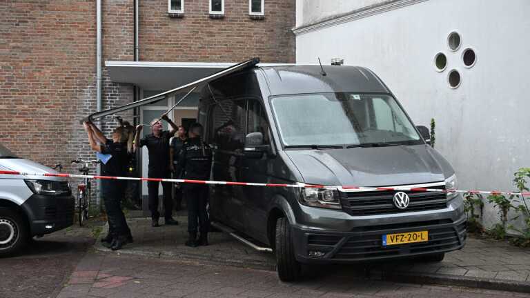 Agenten bij een afgezet gebied tillen een grote plaat van een grijze bestelbus naast een gebouw op