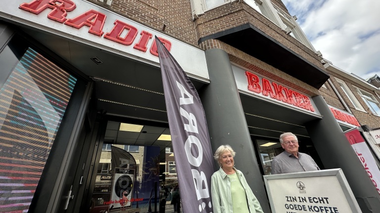 Gevel van Bakkerij Radio met neonletters, zwarte BORRELS-vlag en twee mensen bij een bord met koffie-aanbieding
