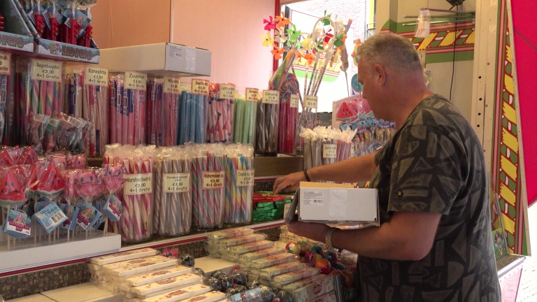 Man bij een snoepkraam met rijen zuurstokken en lollies met prijskaartjes, terwijl hij een doos vasthoudt