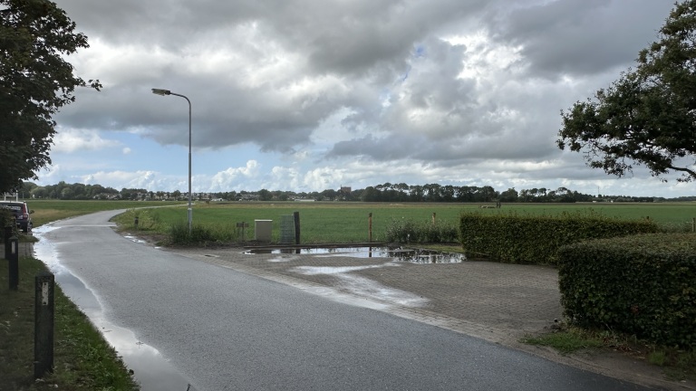 Landweg langs groene weilanden met plassen na regen, lantaarnpaal en zware wolkenlucht boven de horizon