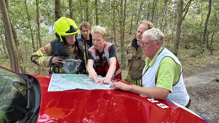 Brandweer en hulpdiensten bespreken een kaart op de motorkap van een rode auto in een bosgebied