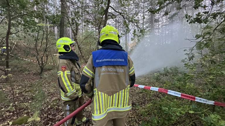 Brandweerlieden in bos blussen brand met brandslang; rook hangt tussen de bomen achter afzetlint