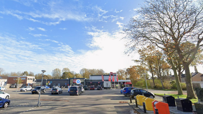 Parkeerterrein met geparkeerde auto’s bij een winkelgebouw, met bomen en afvalcontainers onder een blauwe lucht