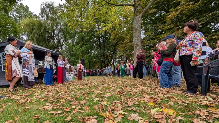 Publiek luistert in een park naar sprekers in traditioneel geborduurde kleding bij een tent, met herfstbladeren op de grond
