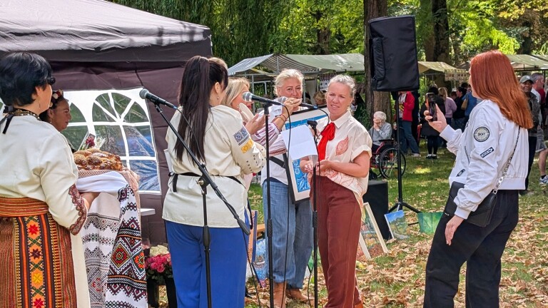 Vrouwen in traditionele kleding staan bij microfoons op een buitenfestival, met publiek en kraampjes op de achtergrond