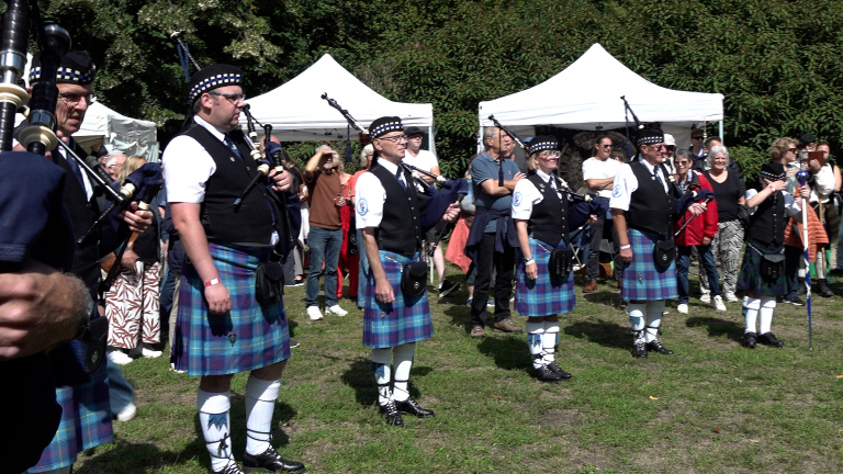 Doedelzakband in kilts speelt buiten op een festival, met publiek en witte tenten op de achtergrond