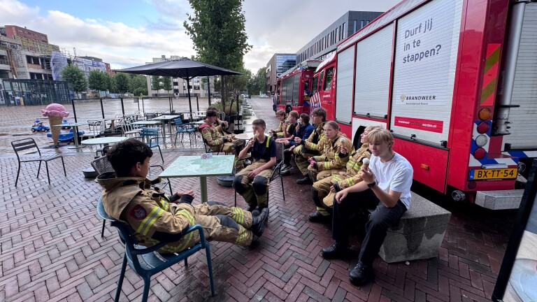Brandweerlieden in pak eten ijs bij een terras naast een rode brandweerwagen met de tekst Durf jij in te stappen?