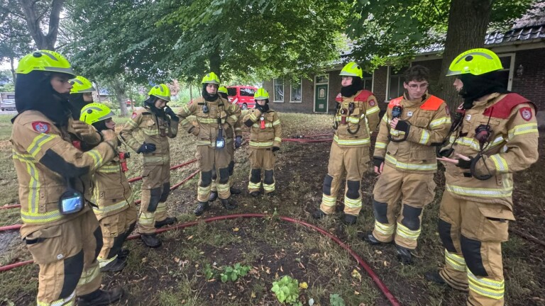 Groep brandweerlieden in uitrukpak en gele helmen luistert buiten bij een gebouw, met brandslangen op de grond