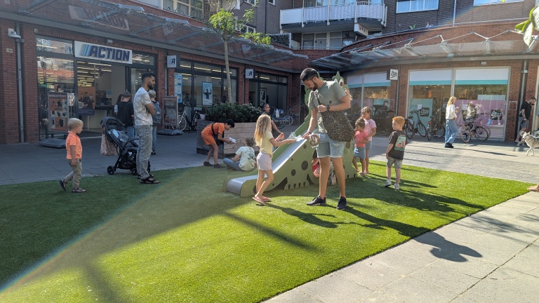 Kinderen spelen op een kleine glijbaan op een grasveldje bij de winkels Action en Etos, met ouders eromheen