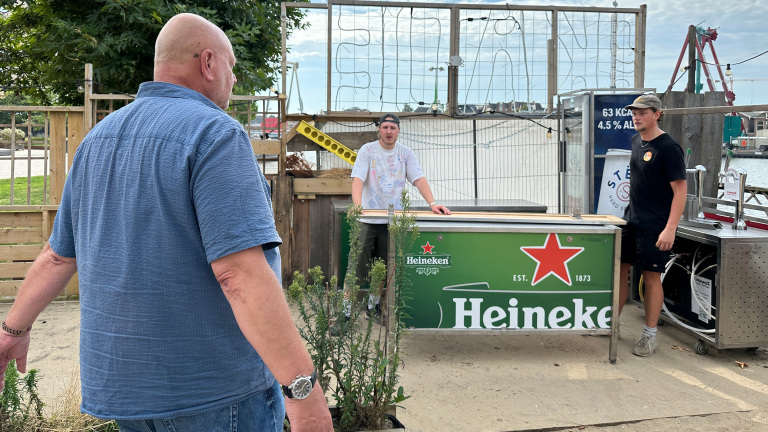 Man in blauw overhemd loopt naar buitenbar met Heineken-balie, waar twee medewerkers bij de tapinstallatie staan