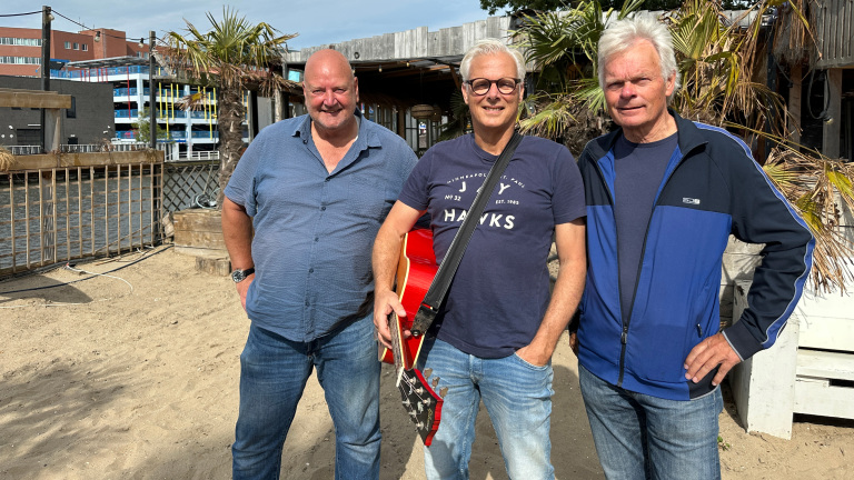 Drie mannen poseren buiten bij een strandpaviljoen met palmbomen; de man in het midden houdt een rode gitaar vast