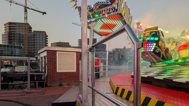 Kermisattractie met felle LED-lichten bij schemering, met bouwkranen en flats op de achtergrond