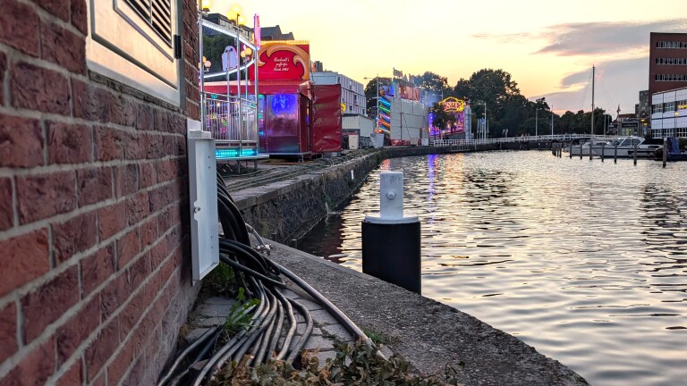 Kermisattracties met gekleurde verlichting langs een kanaal bij zonsondergang, met bakstenen muur en kabels op de kade