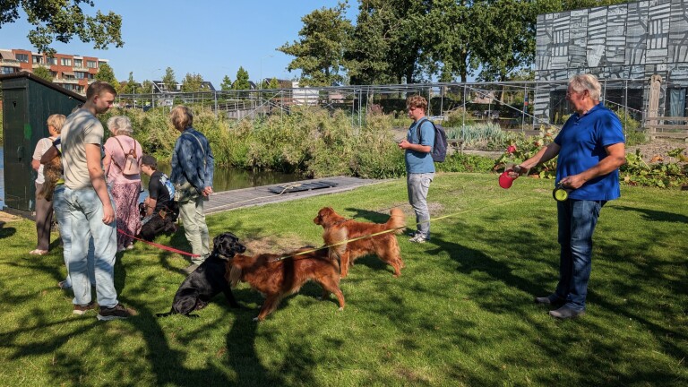 Groep mensen met honden aan de lijn op een grasveld bij een sloot en moestuin, op een zonnige dag