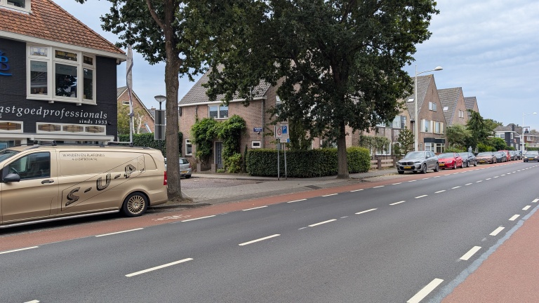 Straat met fietsstrook, geparkeerde auto’s en bestelbus, met bomen en bakstenen huizen langs de weg