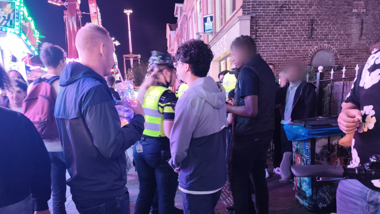 Drukke kermisstraat ’s avonds met politie in gele hesjes die met bezoekers praten bij neonverlichte attracties