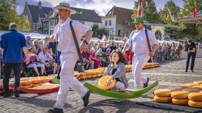 Kaasdragers in wit dragen een brancard met vrouw en kaaswiel, met publiek op een plein vol kaaswielen