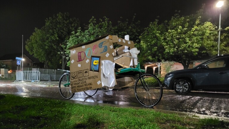 Bakfiets met kartonnen huisje erop, met de tekst “DOOS”, staat ’s nachts in de regen langs de weg