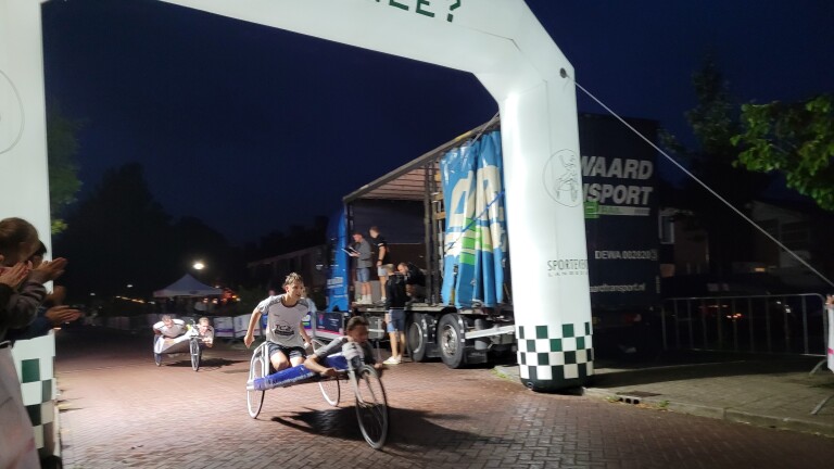 Twee handbikers finishen ’s avonds onder een opblaasbare finishboog, terwijl toeschouwers langs de weg applaudisseren