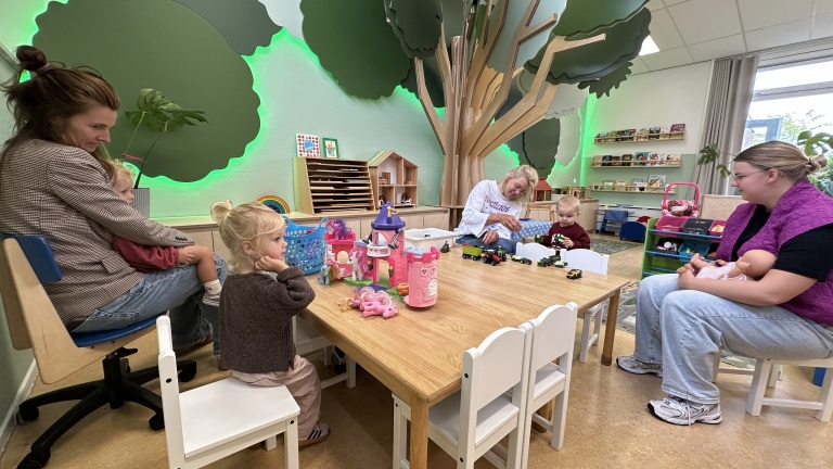 Kinderen spelen met speelgoed op een tafel in een crèche, terwijl drie volwassenen toekijken in een lokaal met muurschildering van bomen