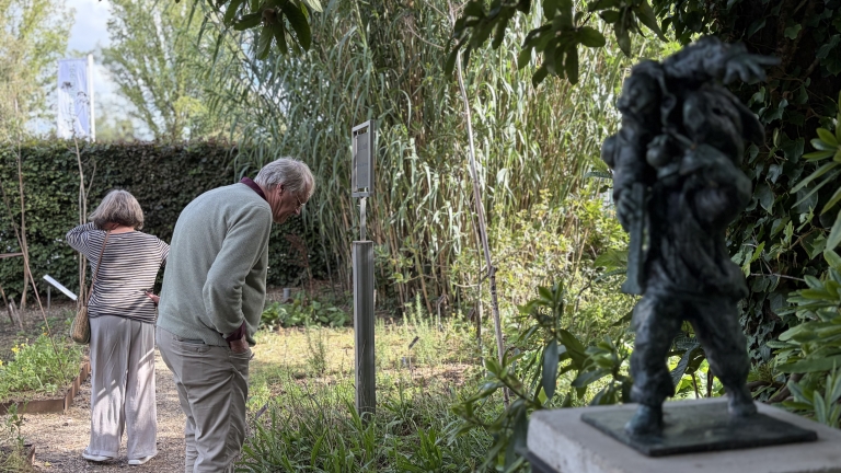 Twee bezoekers bekijken informatieborden in een groene tuin, met rechts op de voorgrond een donker bronzen beeld