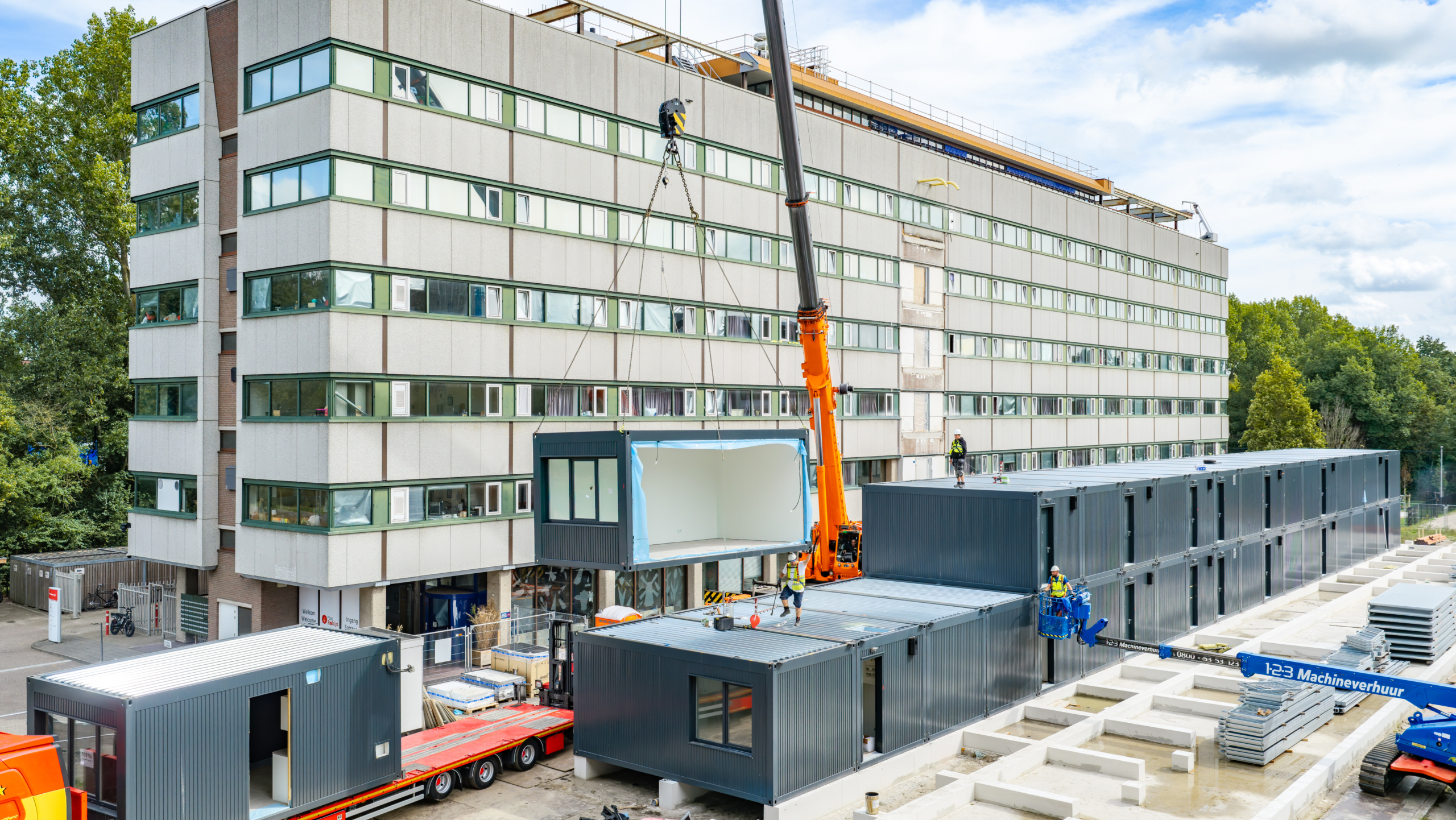 Kraan plaatst een prefab bouwmodule bij een groot kantoorgebouw, met bouwvakkers en stapels grijze containerunits