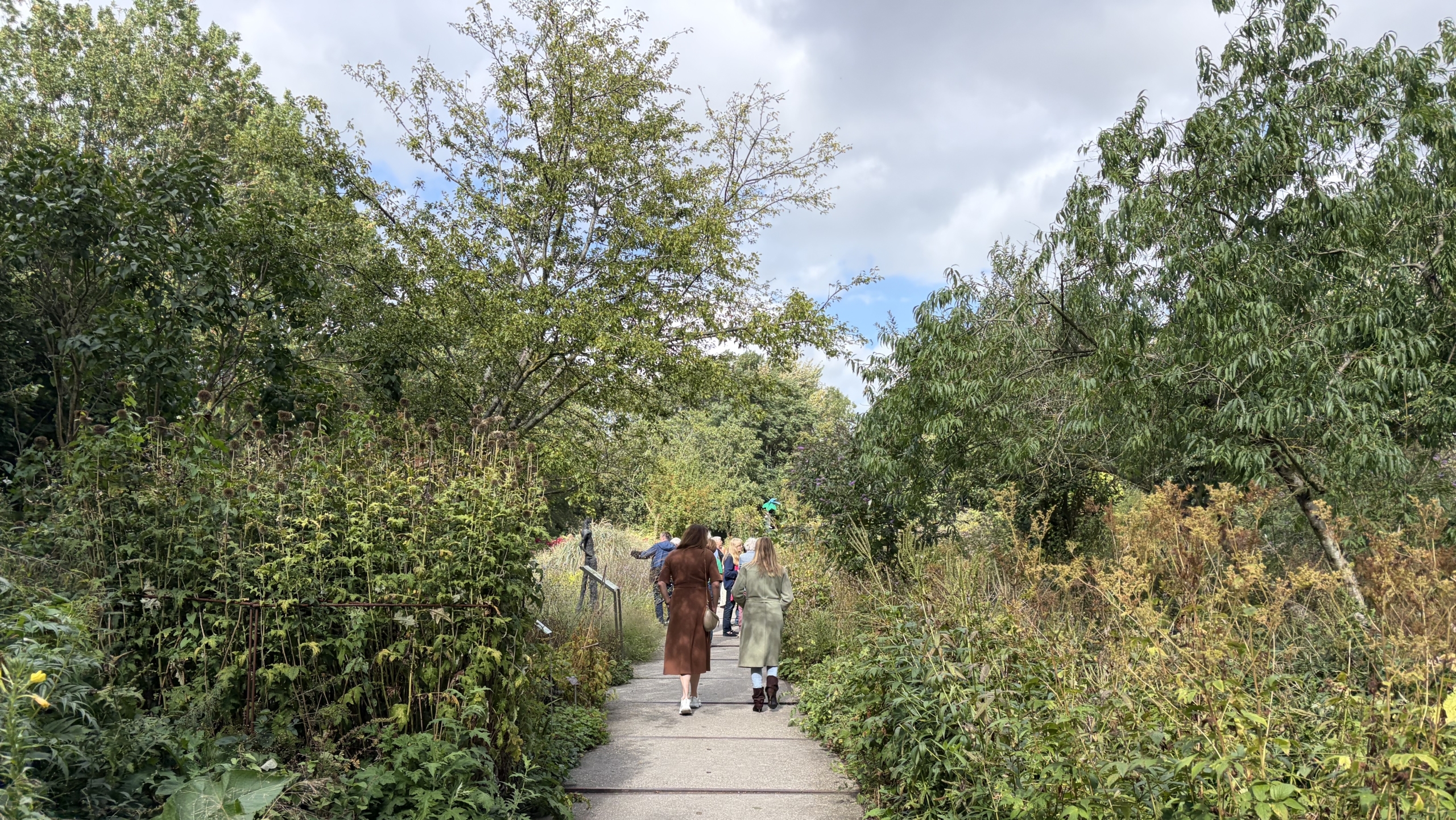 Mensen wandelen over een betonnen pad door een weelderige, groene tuin met hoge planten en bomen onder bewolkte lucht