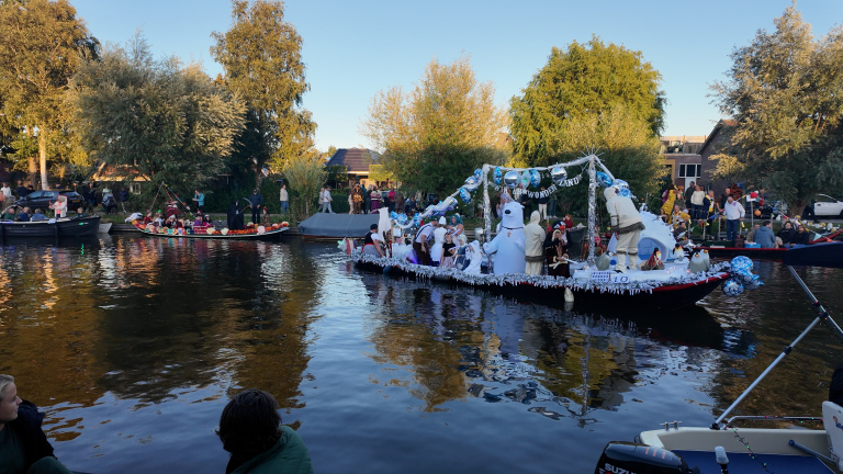 Versierde bootparade op een rivier, met winterthema, sneeuwdecor en verklede deelnemers, bekeken door publiek op de oever