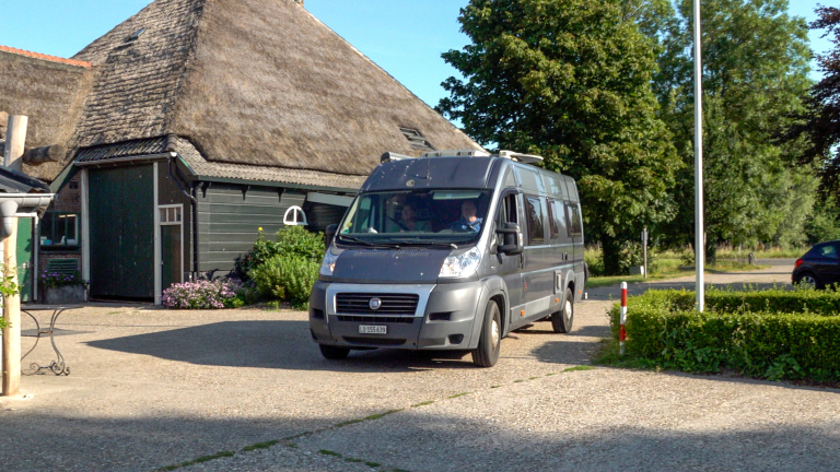 Grijze camperbus geparkeerd voor een boerderij met rieten dak, met bomen en groen op de achtergrond