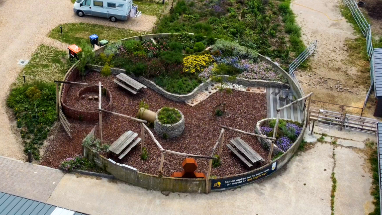 Luchtfoto van omheinde tuin met bloemperken, houtsnipperpad, picknicktafels en verhoogde plantenbakken