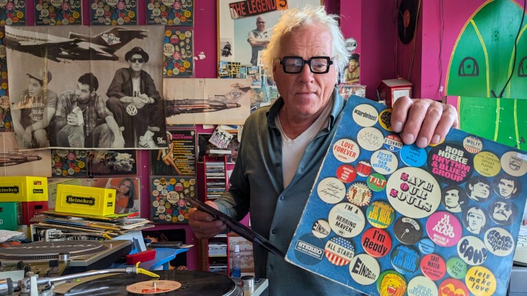 Man met grijs haar en bril houdt sticker-volle platenhoes vast in een platenwinkel met posters en draaitafel