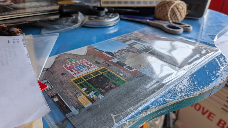 Foto van een gevel met het bord “COOL TIMES”, in een plastic map op een blauwe tafel met schaar en touwbol