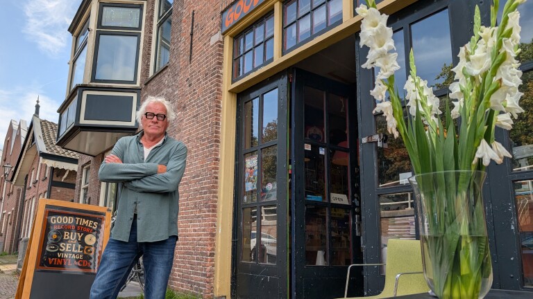 Man met grijs haar en bril staat met gekruiste armen voor de ingang van platenzaak Good Times, met witte bloemen op voorgrond