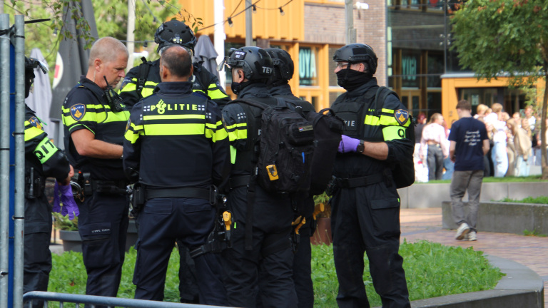 Groep Nederlandse politieagenten in uniform en helmen overlegt op straat, met omstanders op de achtergrond