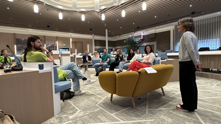 Vrouw spreekt een groep toe in een raadszaal; mensen zitten op banken en stoelen en luisteren naar haar