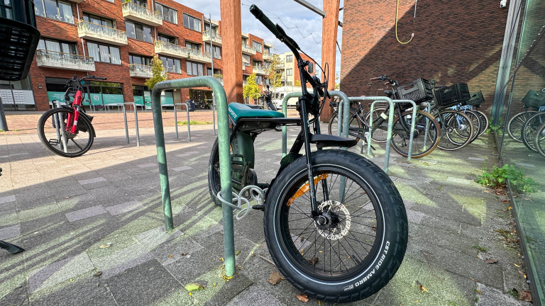 Zwarte fatbike met brede voorband vast aan fietsenrek, met andere fietsen en bakstenen appartementen op de achtergrond
