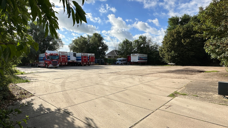 Groot betonnen parkeerterrein met geparkeerde vrachtwagens en een camper, omringd door bomen onder een blauwe lucht