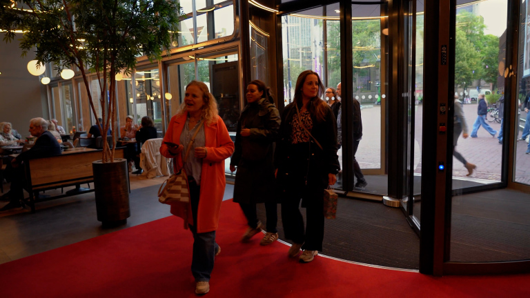 Vrouwen lopen over een rode loper een modern gebouw binnen via een draaideur, met zitgedeelte en grote ramen