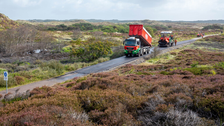 Een vrachtwagen met een rode kiepbak stort materiaal uit op een smalle weg in een groen heideachtig landschap, terwijl een asfaltmachine het materiaal verwerkt. Arbeiders zijn aan het werk langs de weg.