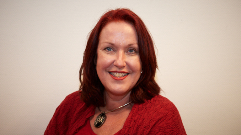 Portret van een glimlachende vrouw met rood haar, rode trui en ketting met hanger tegen een lichte achtergrond